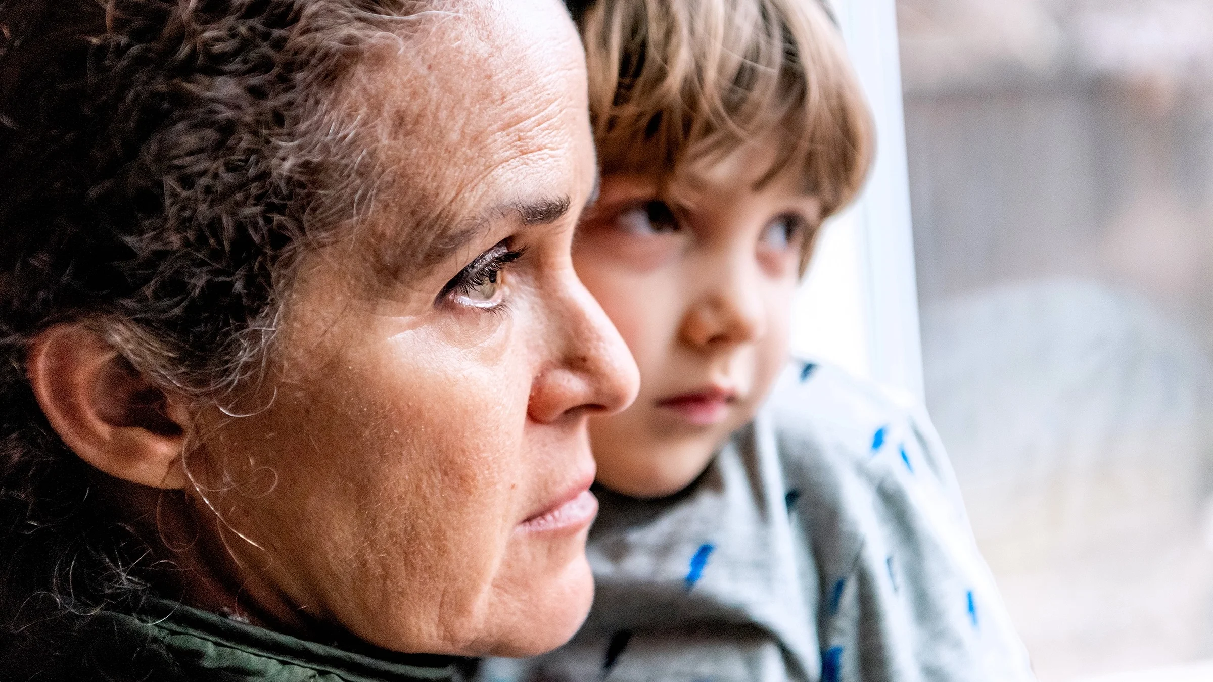 Close-up portrait of a woman and her son. They are looking out a window to the street and both have sad but somewhat neutral expressions.