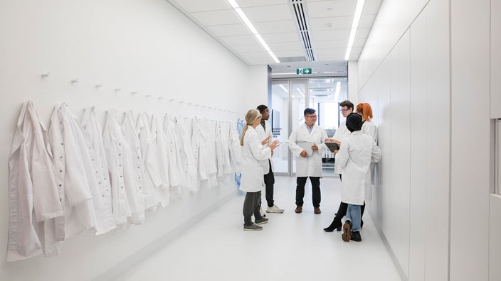 Doctors in white coats circled up, going over the schedule. They are standing in a very long, white hospital hallway.