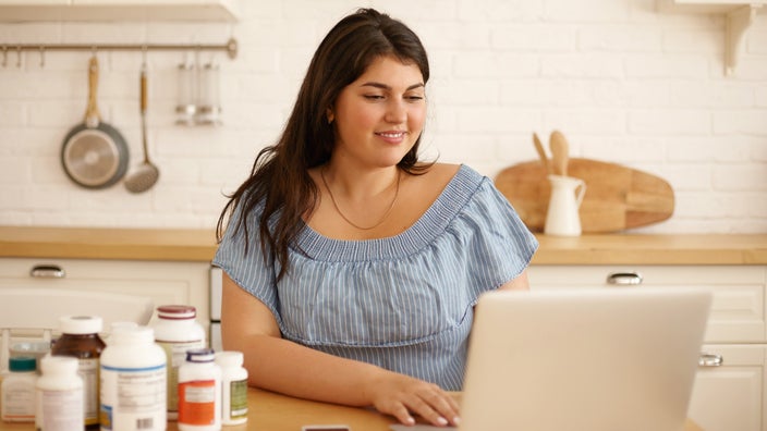 Plus size woman sitting at her laptop in the kitchen surrounded by supplement bottles on the table.