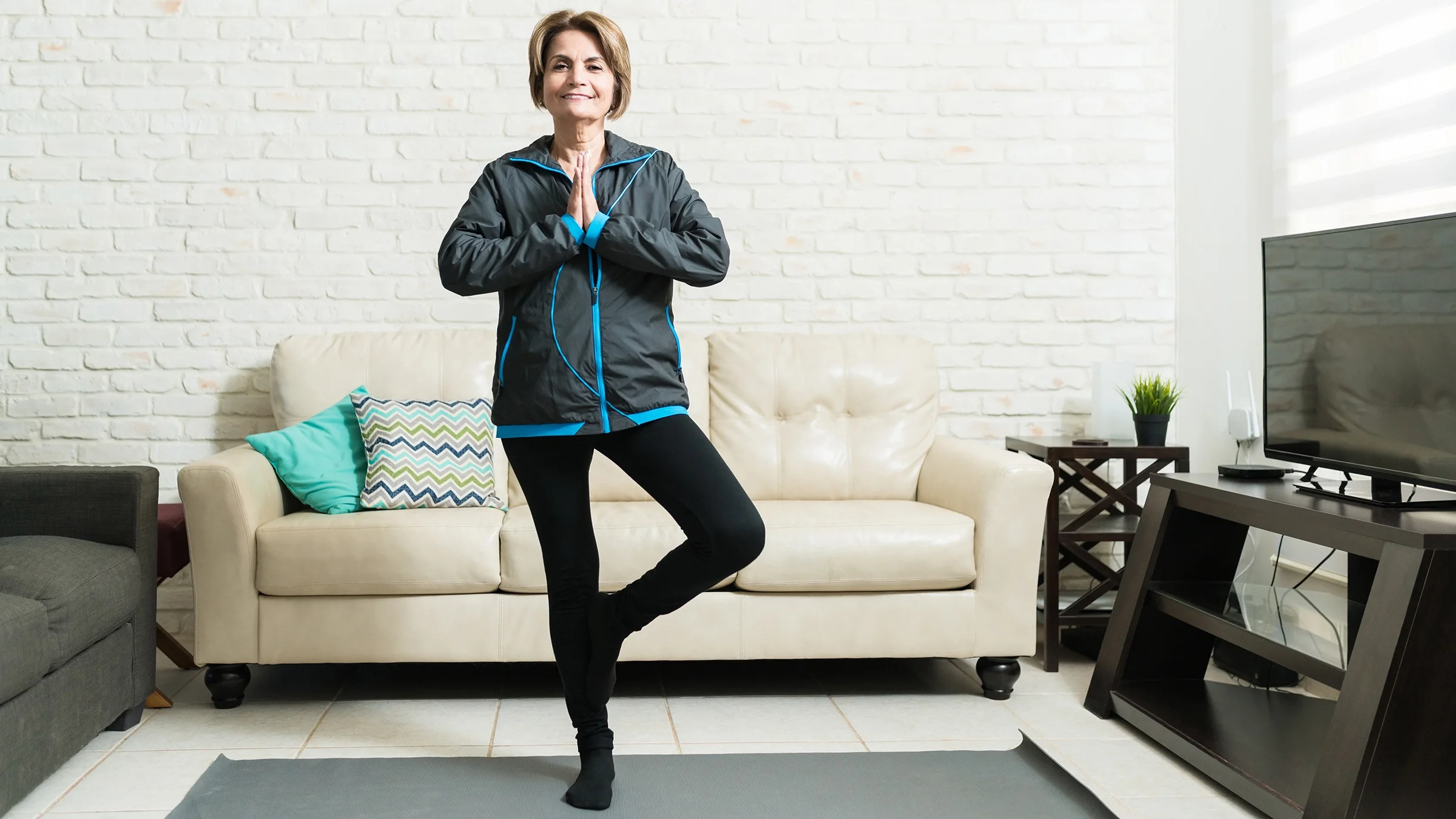 Senior woman practices a tree pose on an exercise mat in her living room.