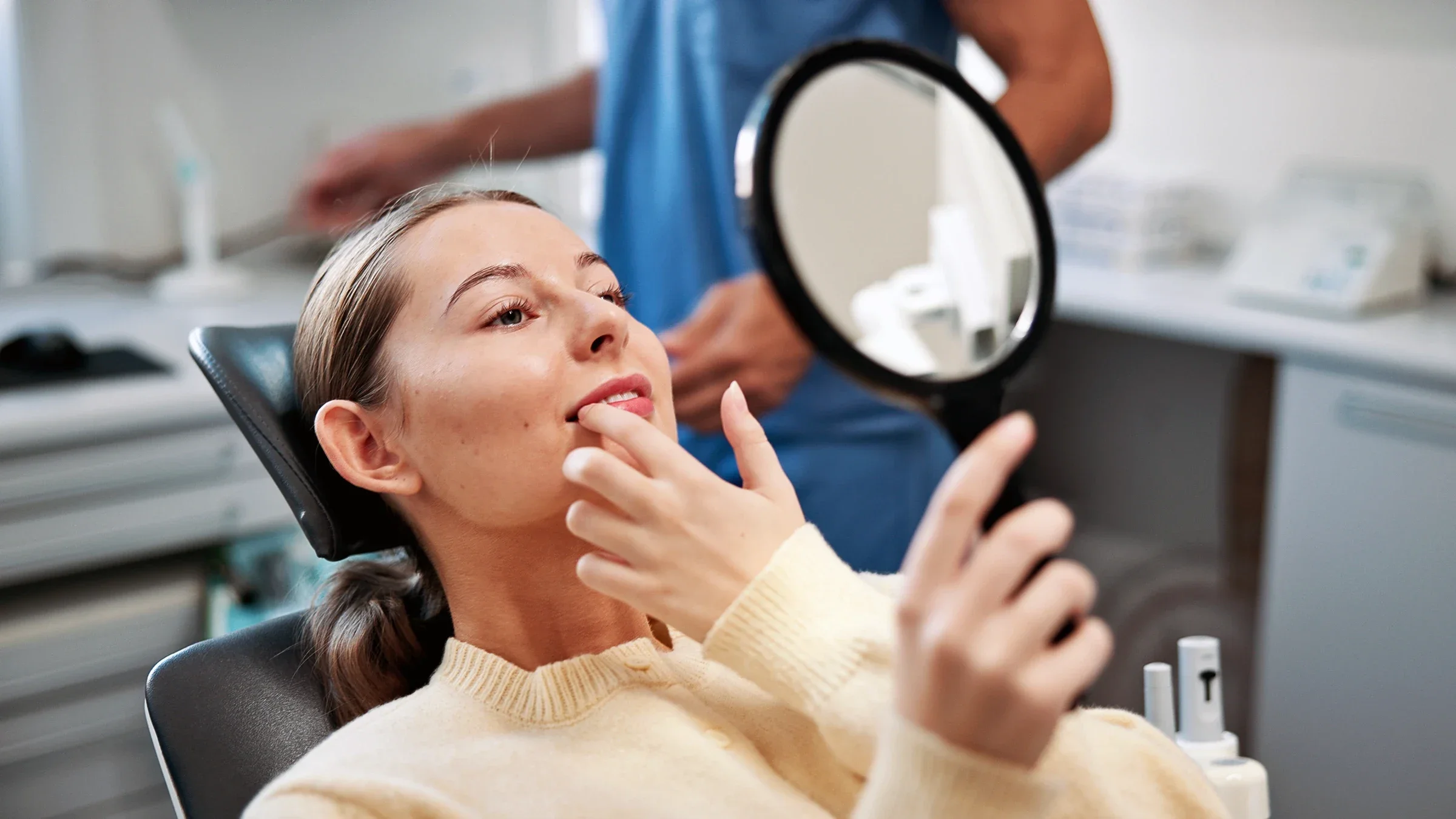 A woman looks at her teeth while holding a mirror during a dental appointment. 
Scandistock/E+ via Getty Images