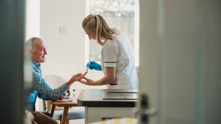 A nurse testing an older man's blood sugar in the background in focus with a doorway in the foreground out of focus.