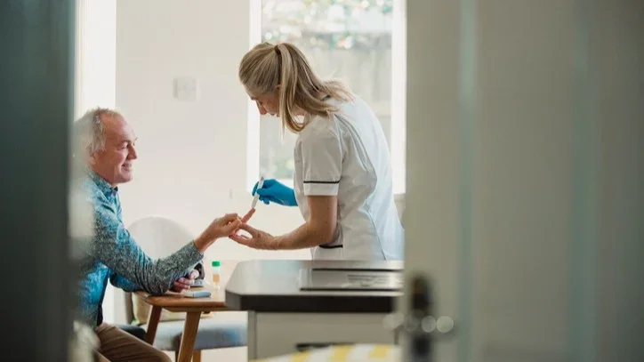 A nurse testing an older man's blood sugar in the background in focus with a doorway in the foreground out of focus.
DGLimages/iStock via Getty Images