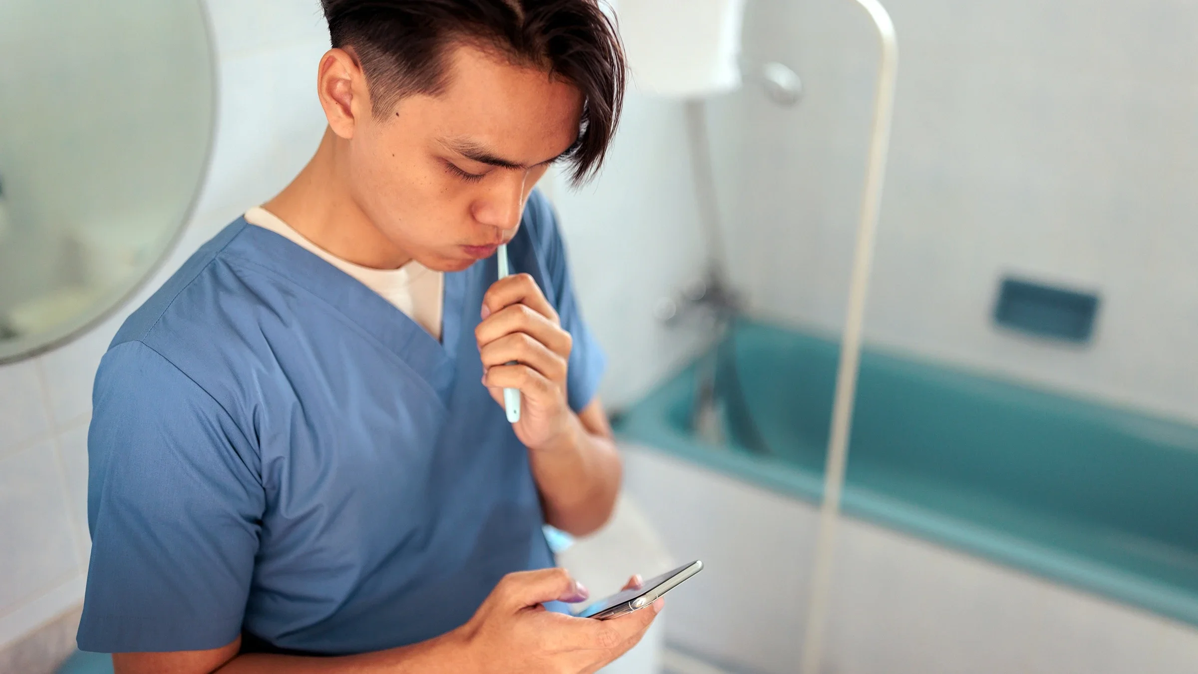 Man wearing scrubs brushing his teeth while checking his phone.