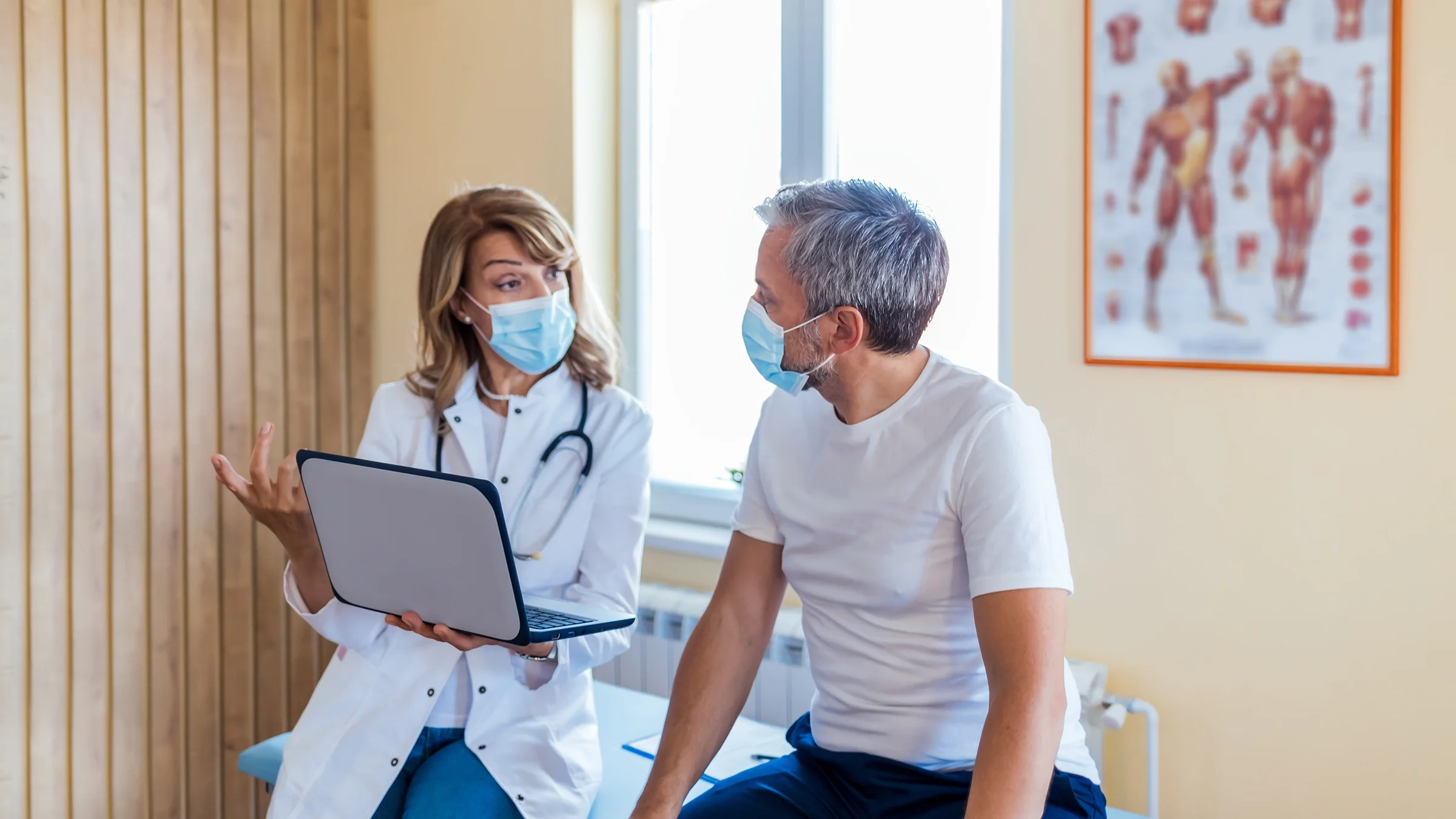 An older adult is having a consultation with his doctor in the exam room. They are both sitting on the exam table wearing face masks.
