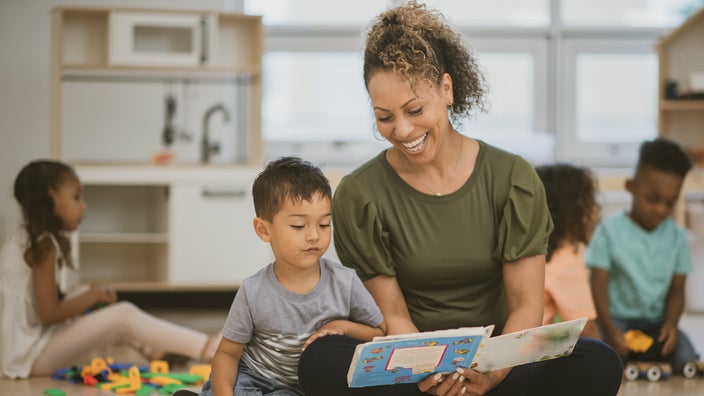 Smiling preschool teacher sitting crosslegged reading to her student. The toddler is leaning on her leg looking at the book.