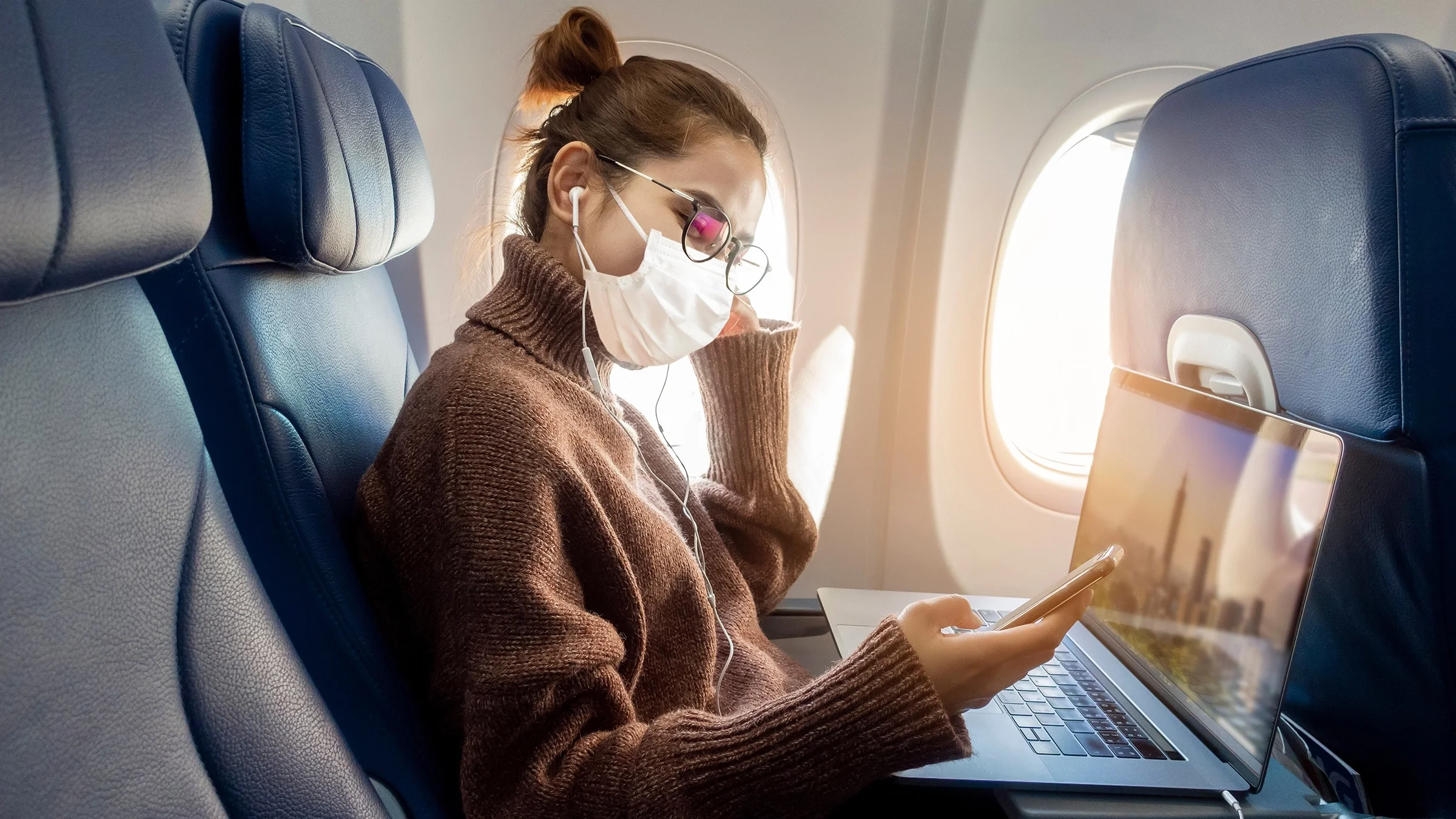 Woman sitting on an airplane with the tray table down with her laptop on it. She is wearing glasses, headphones, and a white face mask.