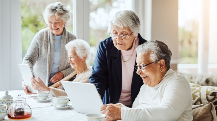 Group of four elderly woman gathered for afternoon tea while looking at information on a tablets, two women are sitting down while two others look over their shoulders.
