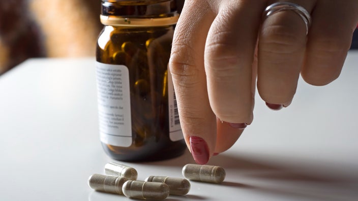 Person with red painted nails picking up a supplement capsule from a pile. The medicine bottle is behind the pile of pills.