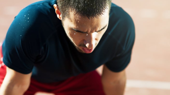 Close-up of a man pausing in his run to take a break. He is leaning over resting his hands on his knees and is dripping in sweat.