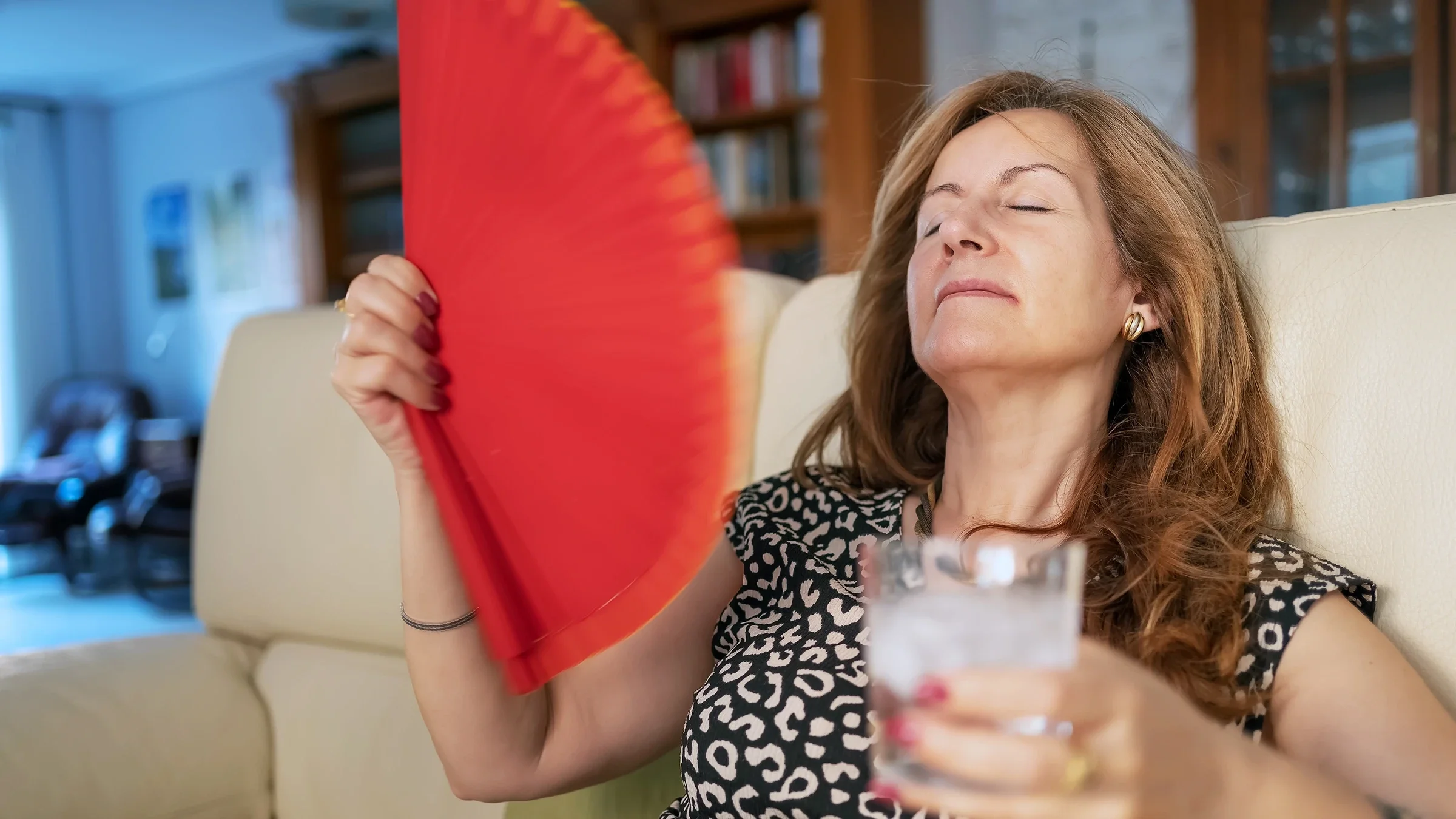 A woman is fanning herself and drinking water at home during a hot flash.