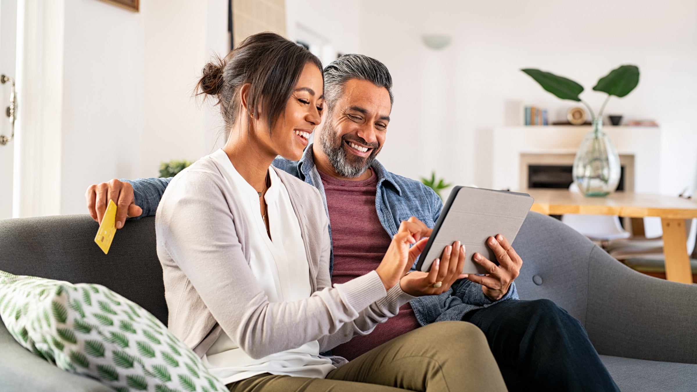 A smiling couple on a couch holding a tablet. The woman is pointing at something on the tablet screen, and the man is holding a credit card in his hand.