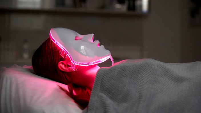 A woman wears a red light mask while lying on a bed.