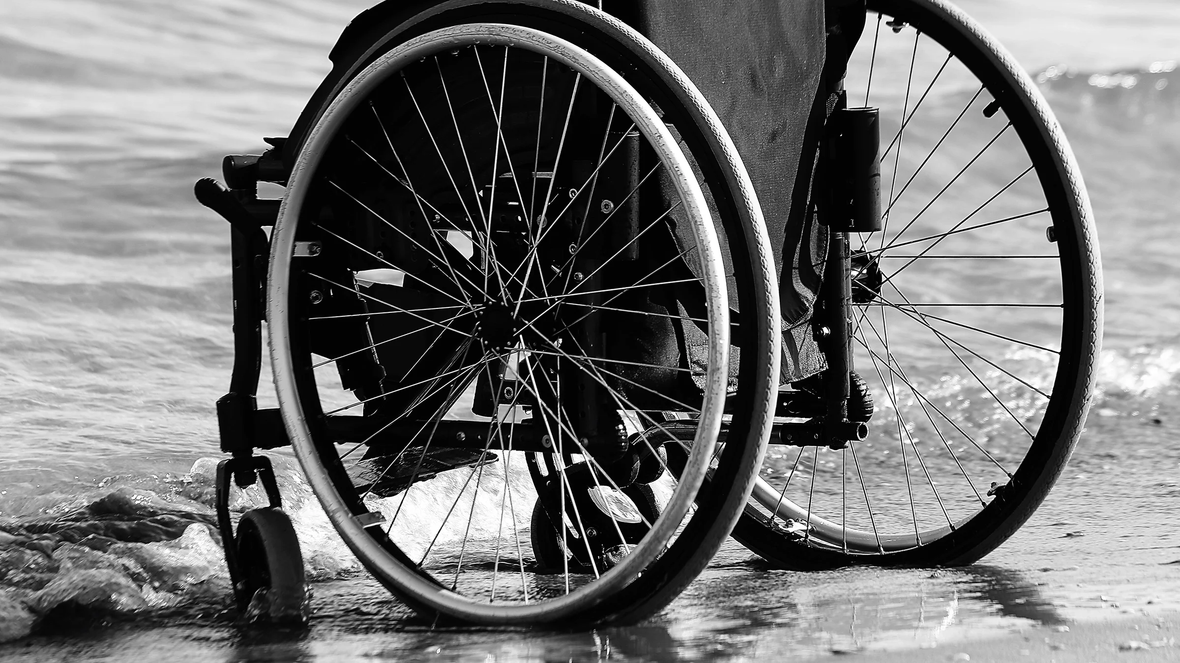A cropped black and white shot of a person in a wheelchair on the beach at the water's edge.