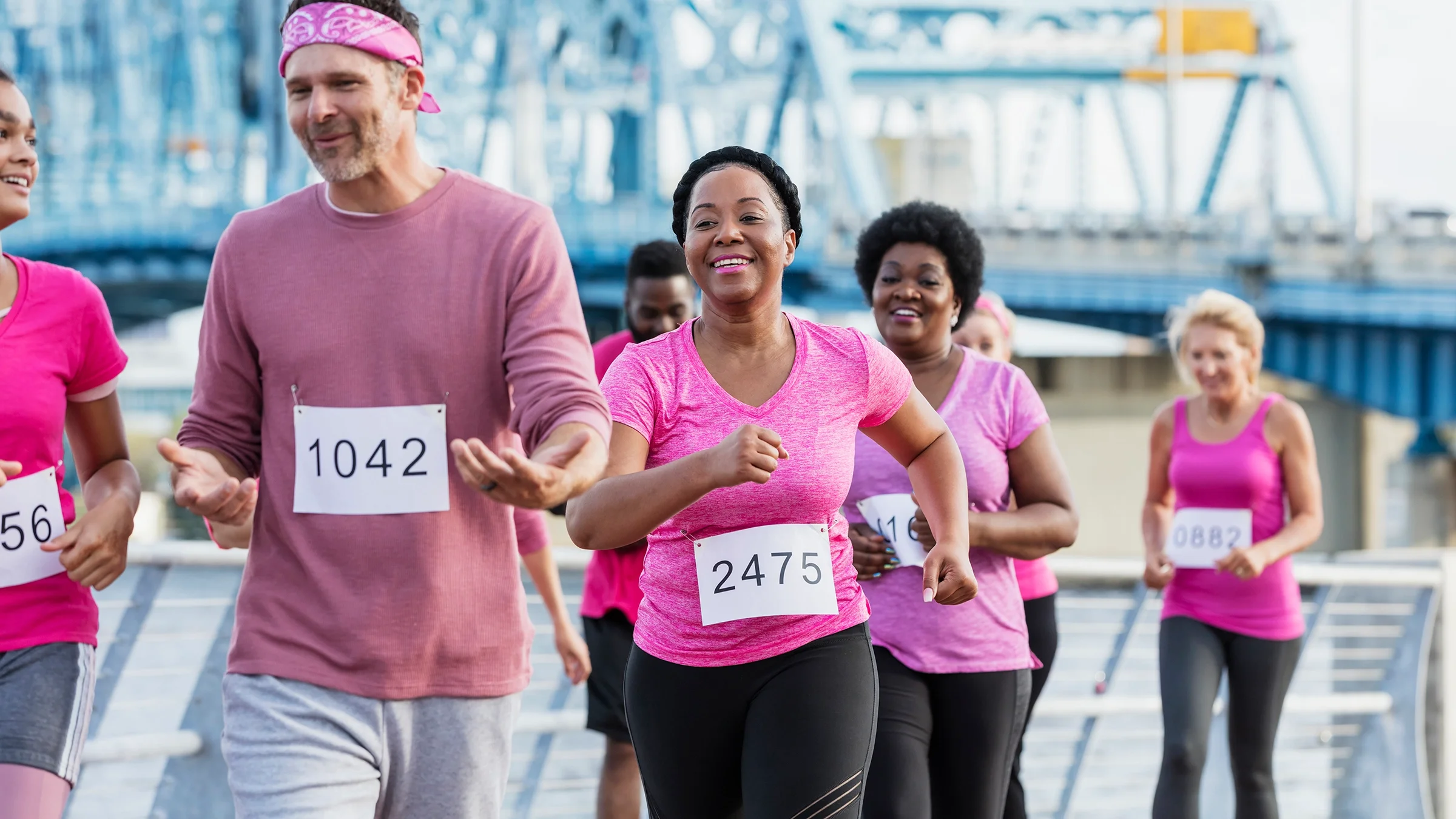 Group of people running for a breast cancer awareness run. They are running along the water next to a bridge.