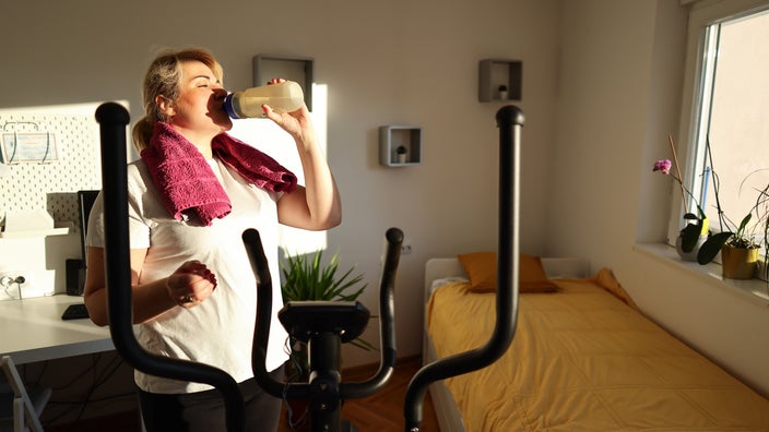A woman drinks water after finishing a workout in her bedroom.