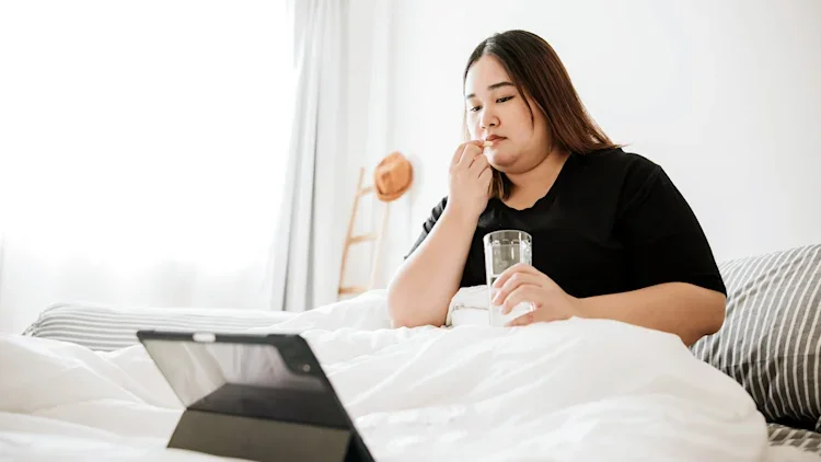A woman sits up in bed watching her tablet and preparing to take a pill with a glass of water.