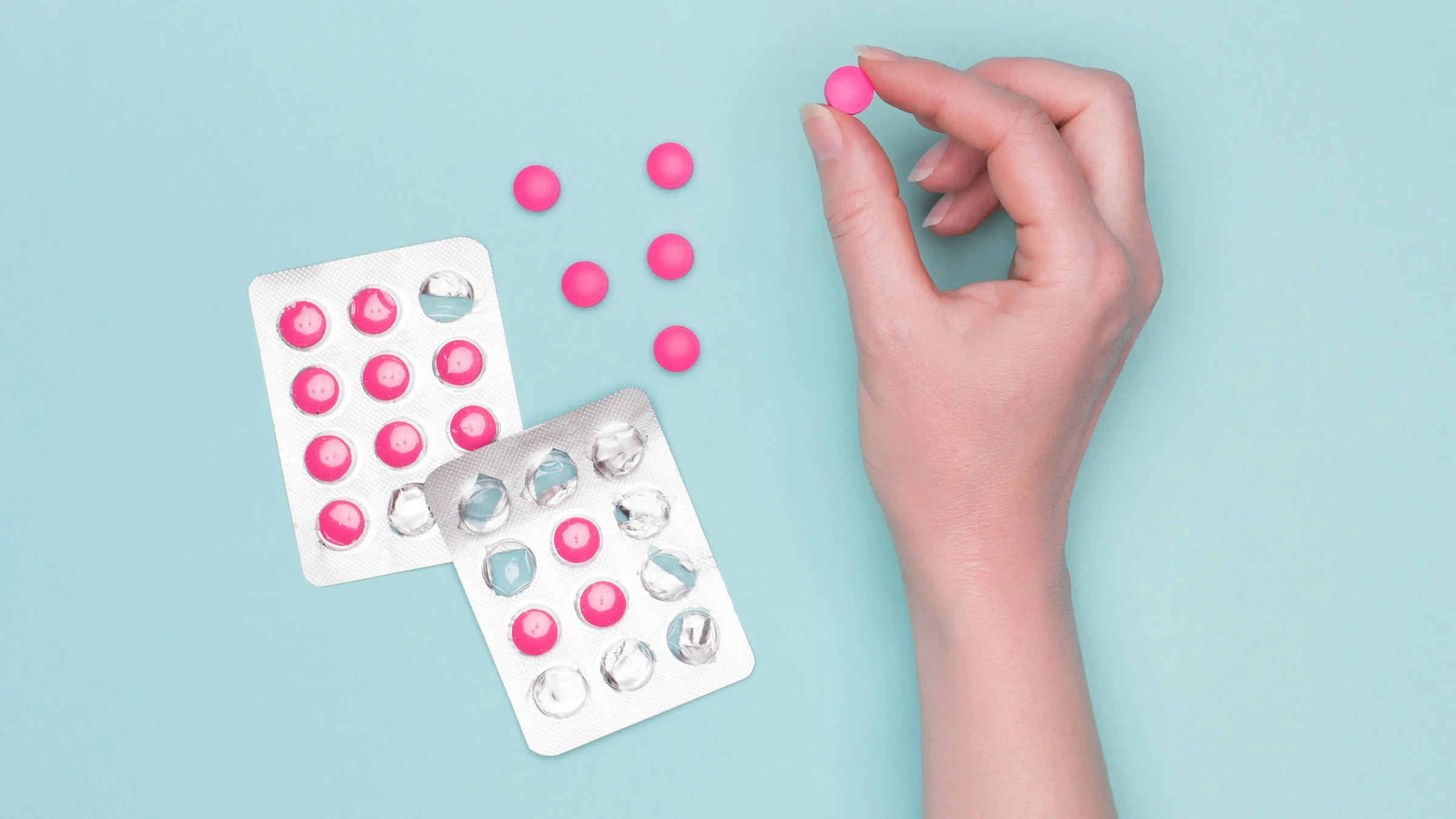 Hand holding one pink Benadryl pill in-between thumb and pointer finger with other pink pills in the blister pack and some on out of the pack on a light blue background.