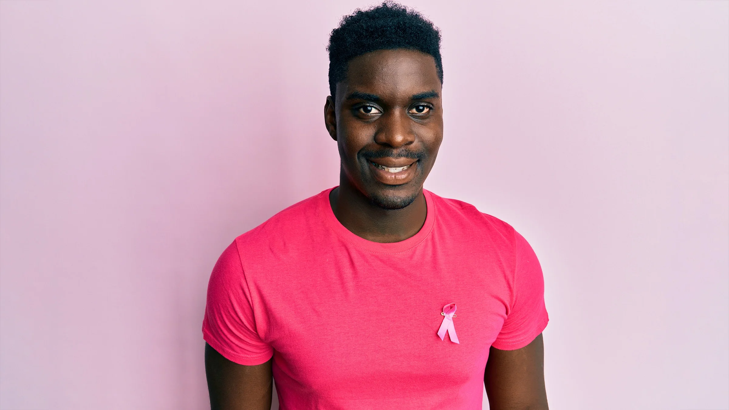 Portrait of a young man wearing a pink t-shirt and pink breast cancer awareness ribbon on it. The background is a plain pink studio backdrop.