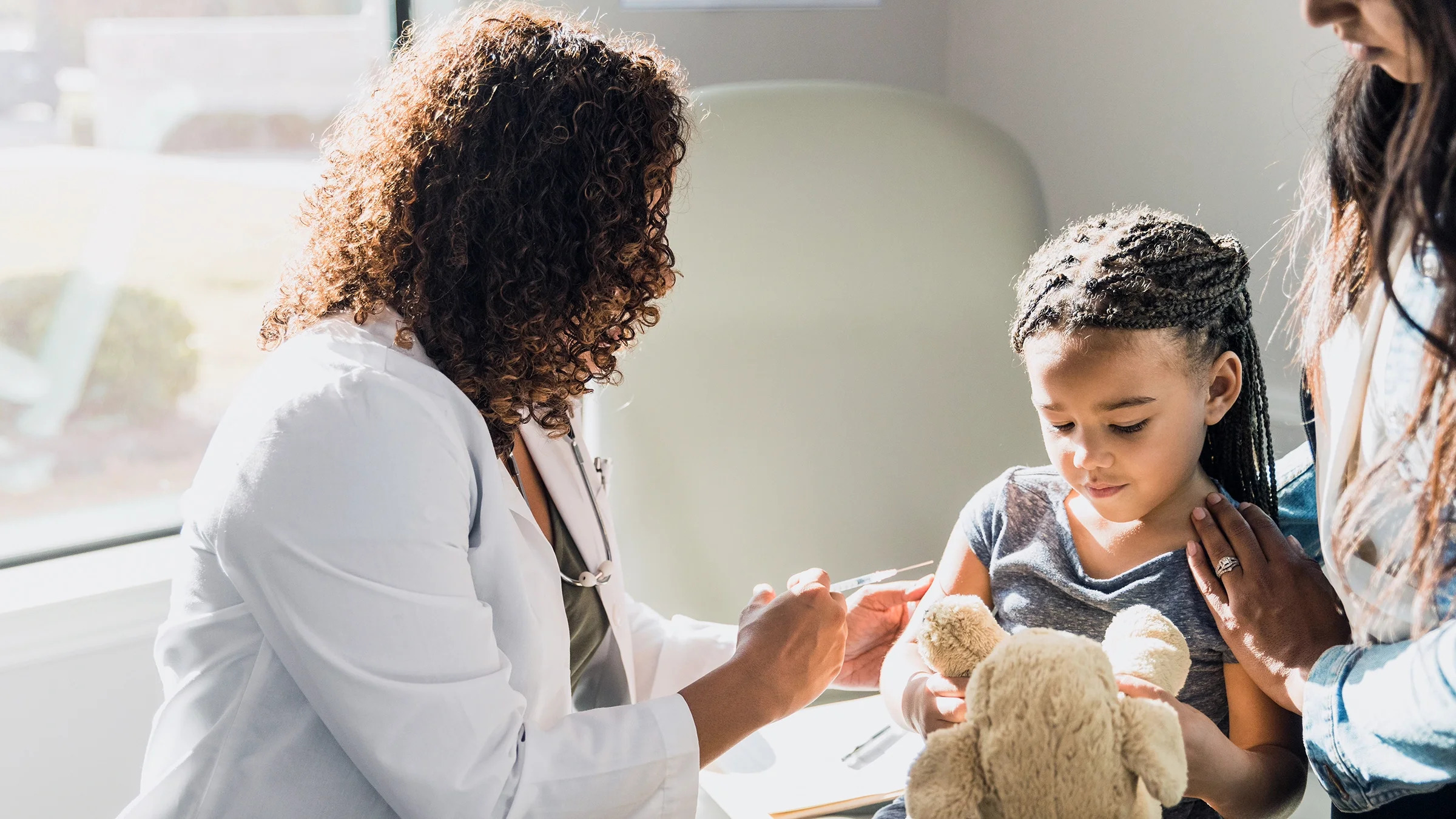 A girl holds a Teddy bear as she gets a vaccine.