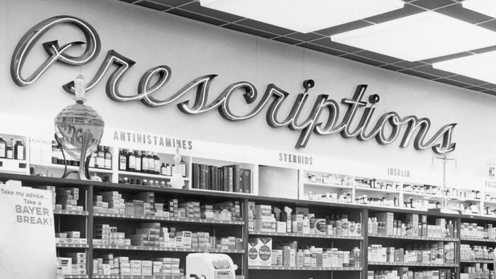 Black and white vintage photo of a 1950s pharmacy. There is a large neon cursive sign reading "Prescriptions" above the shelves of medication.