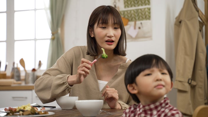 A mom trying to feed broccoli to her child, who is turning away.