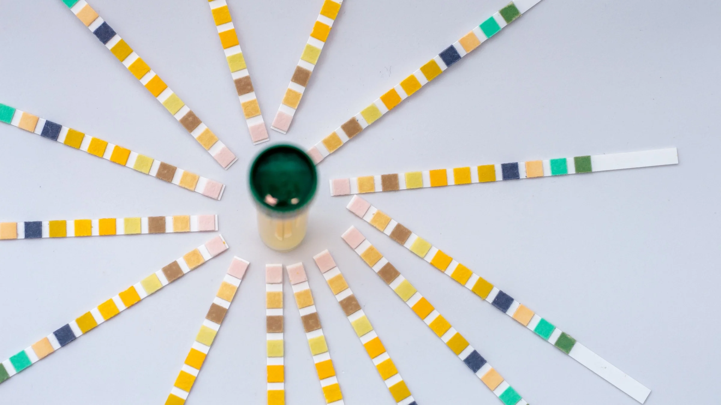 Flat lay image of a urine sample with test strips with different colored squares radiate off of the sample like a sunburst.