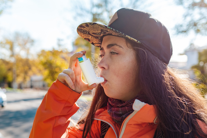 Close-up portrait of a young woman in a baseball cap using an inhaler outside.