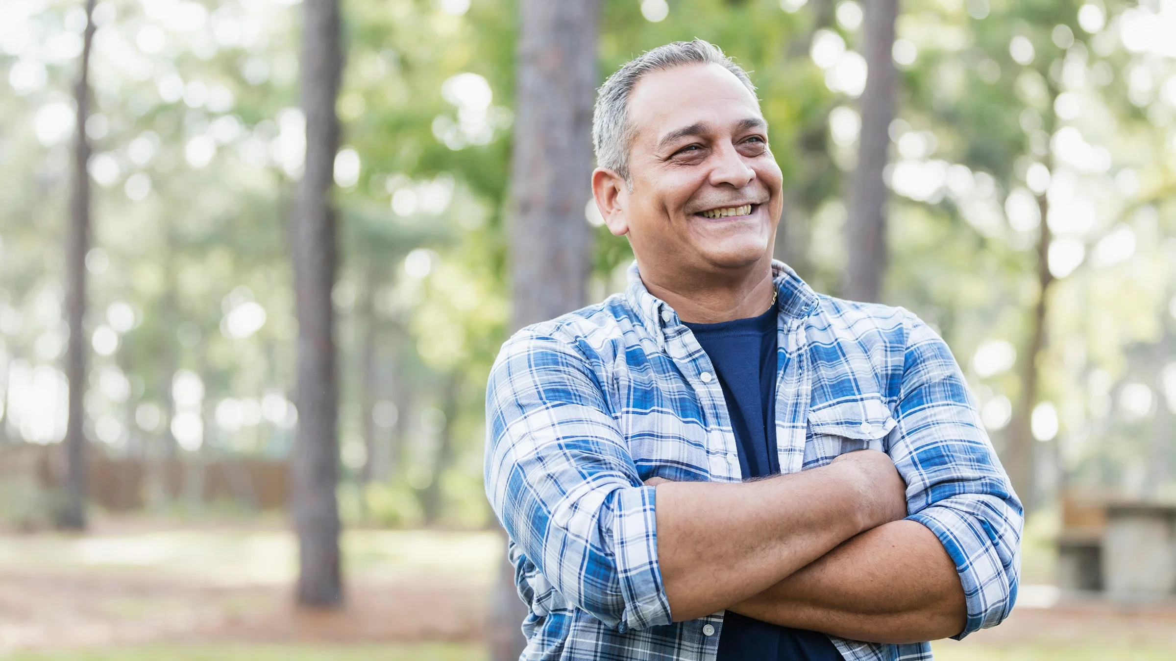 Portrait of a smiling man in the woods. He is wearing a blue and white plaid shirt.