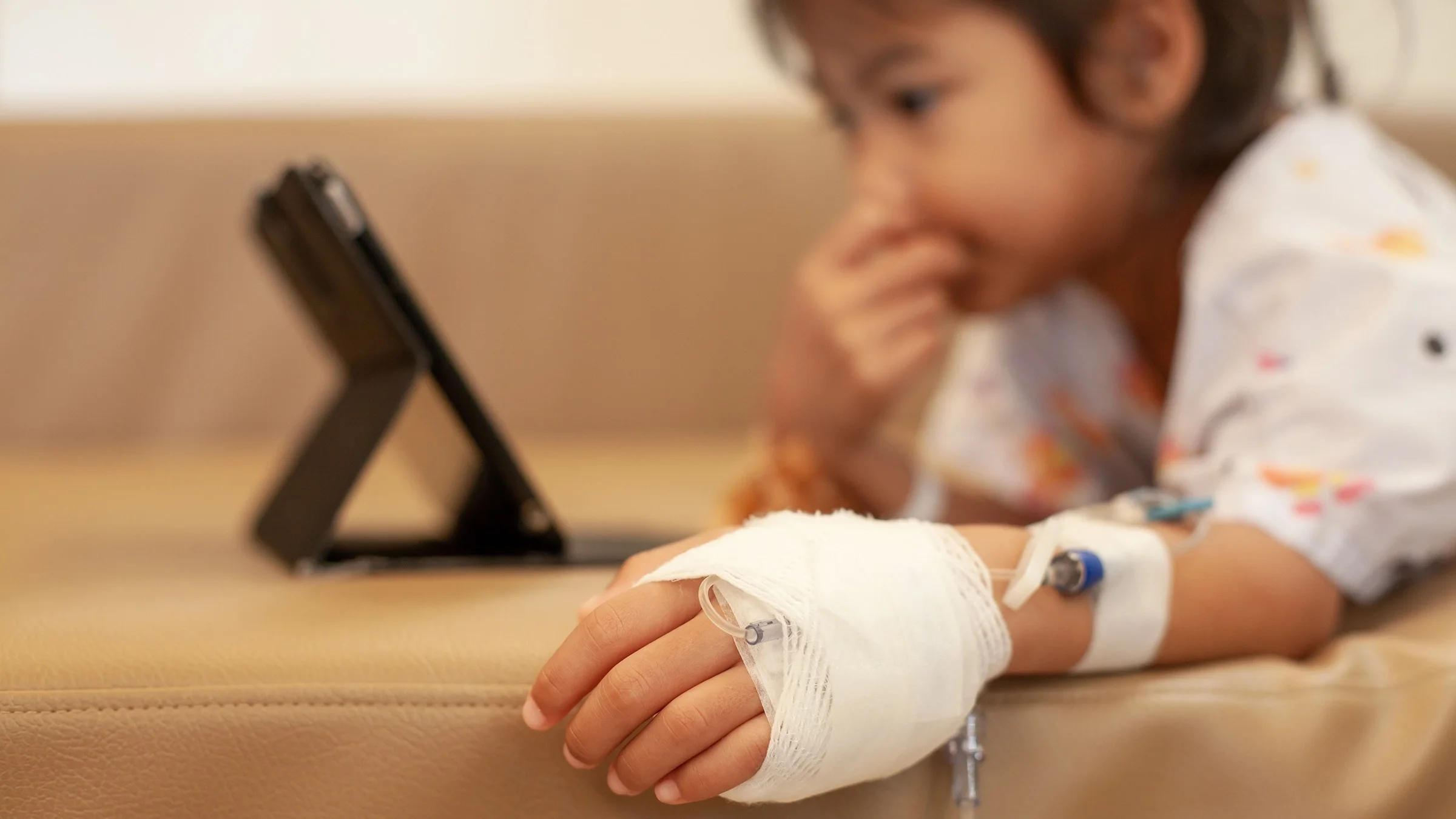 Close-up on a little girl's hand with an IV bandaged to it. She is in the background out of focus watching something on a tablet.