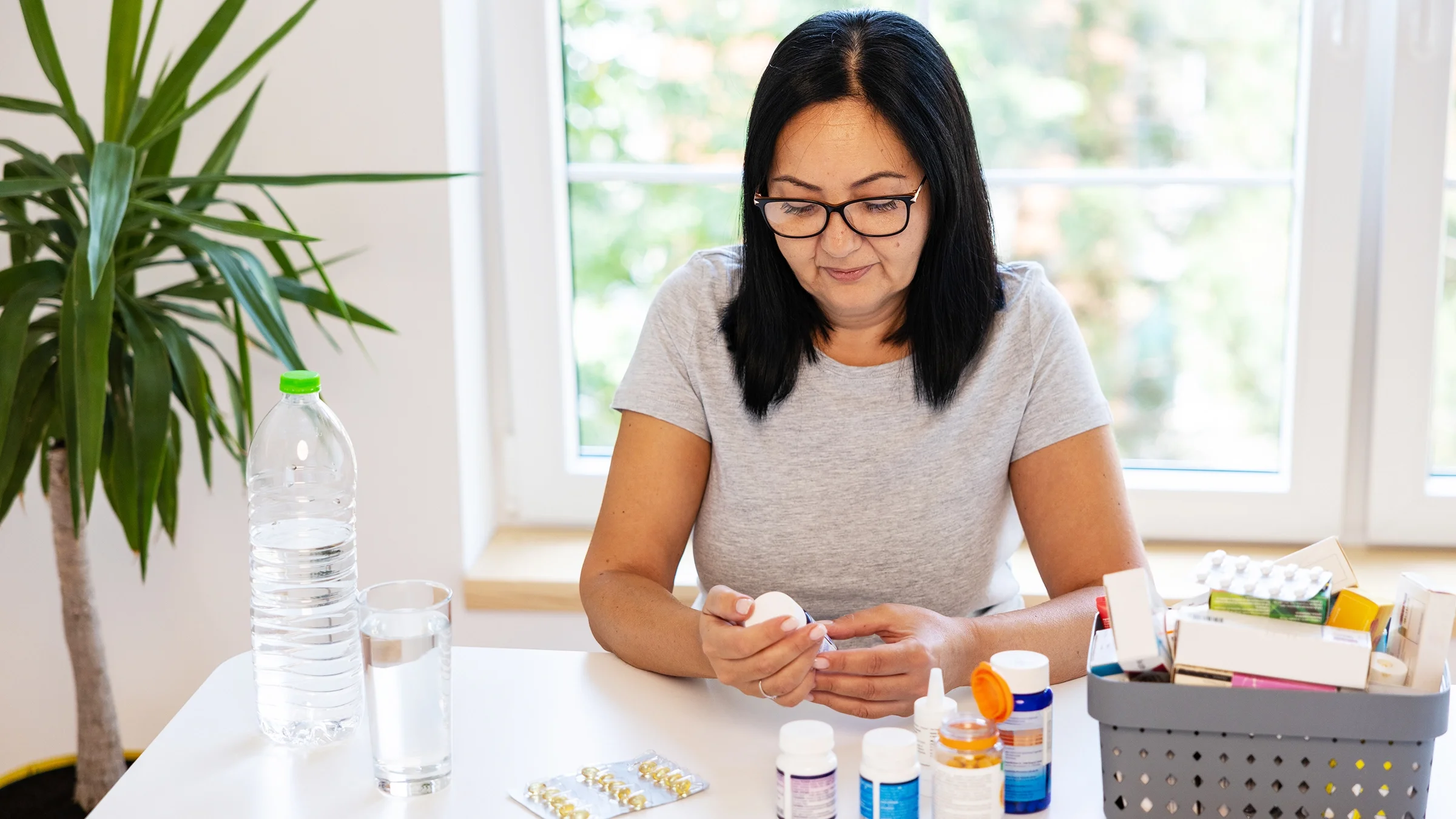 A woman reads the label on a pill bottle while sitting at a dining table.