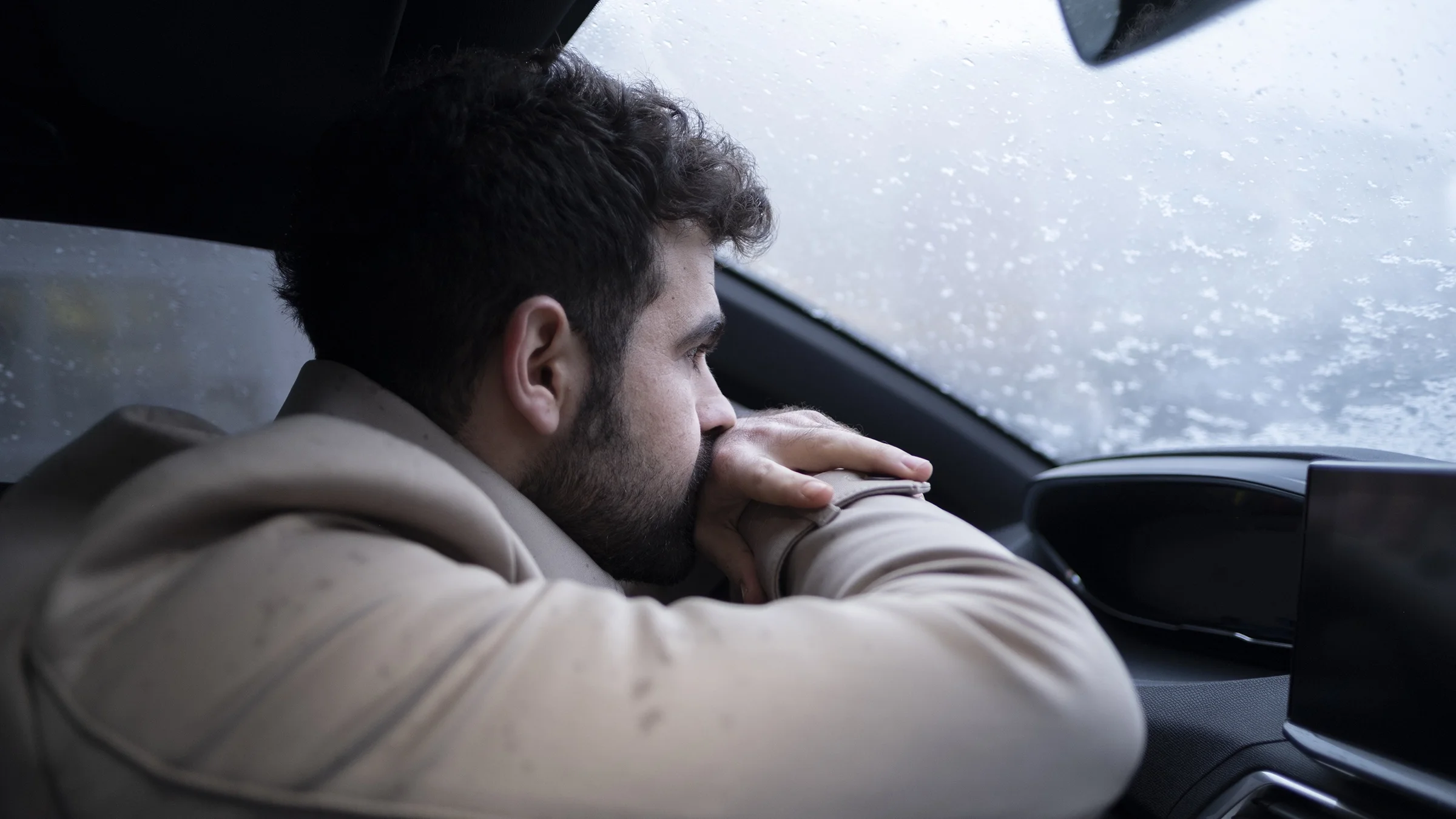 A man experiencing sadness leans against the steering wheel of his vehicle with an overwhelmed expression.