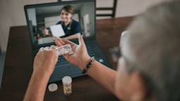 Close-up of an elderly man at his laptop for a telehealth visit showing his nurse the medication he has in hand.
Edwin Tan/E+ via Getty Images