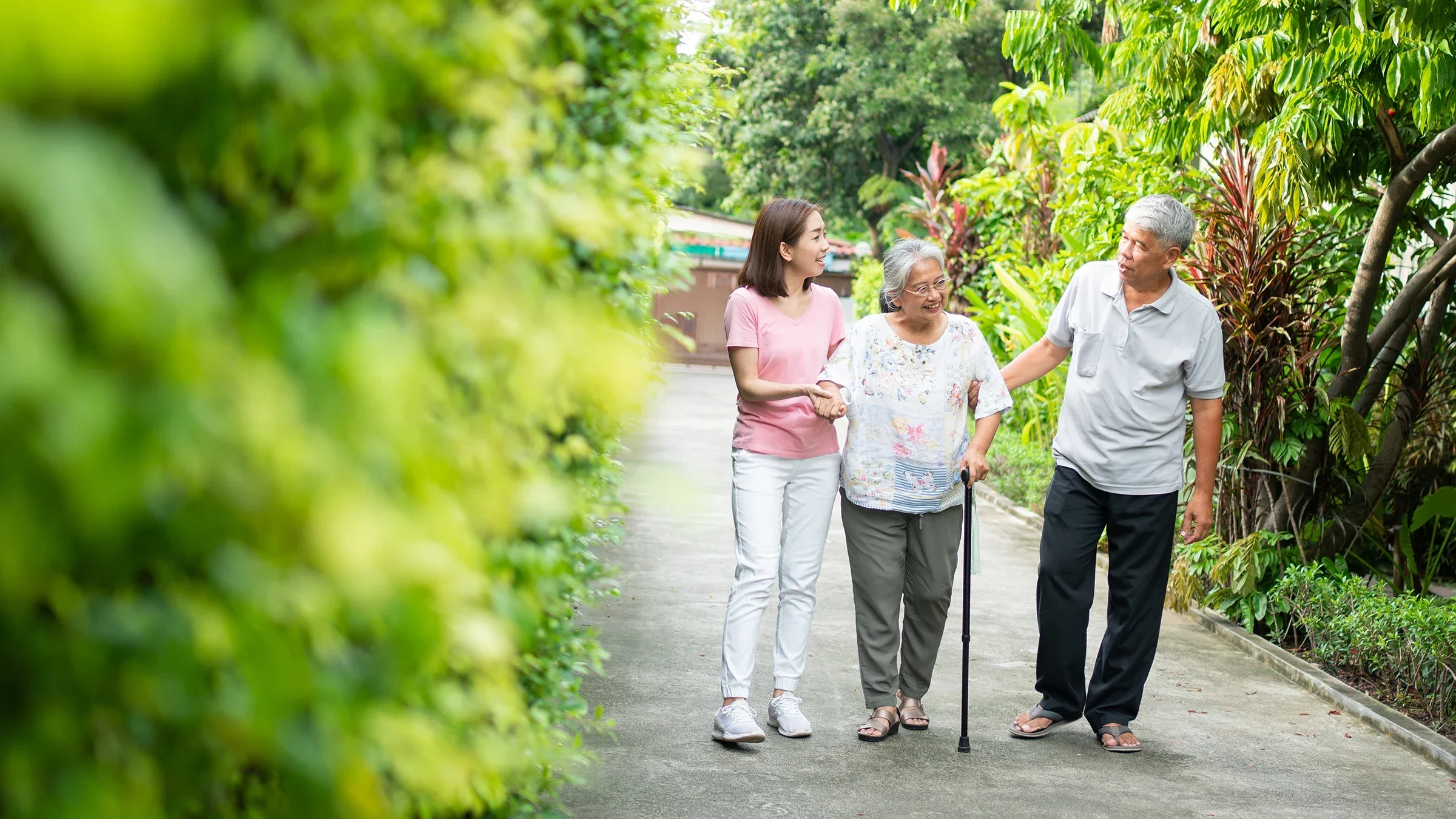 Adult daughter helping her mother on a family walk down a lush and green pathway. The mother is using a cane to walk and the father is on the other side of her.
Prot Tachapanit/iStock via Getty Images