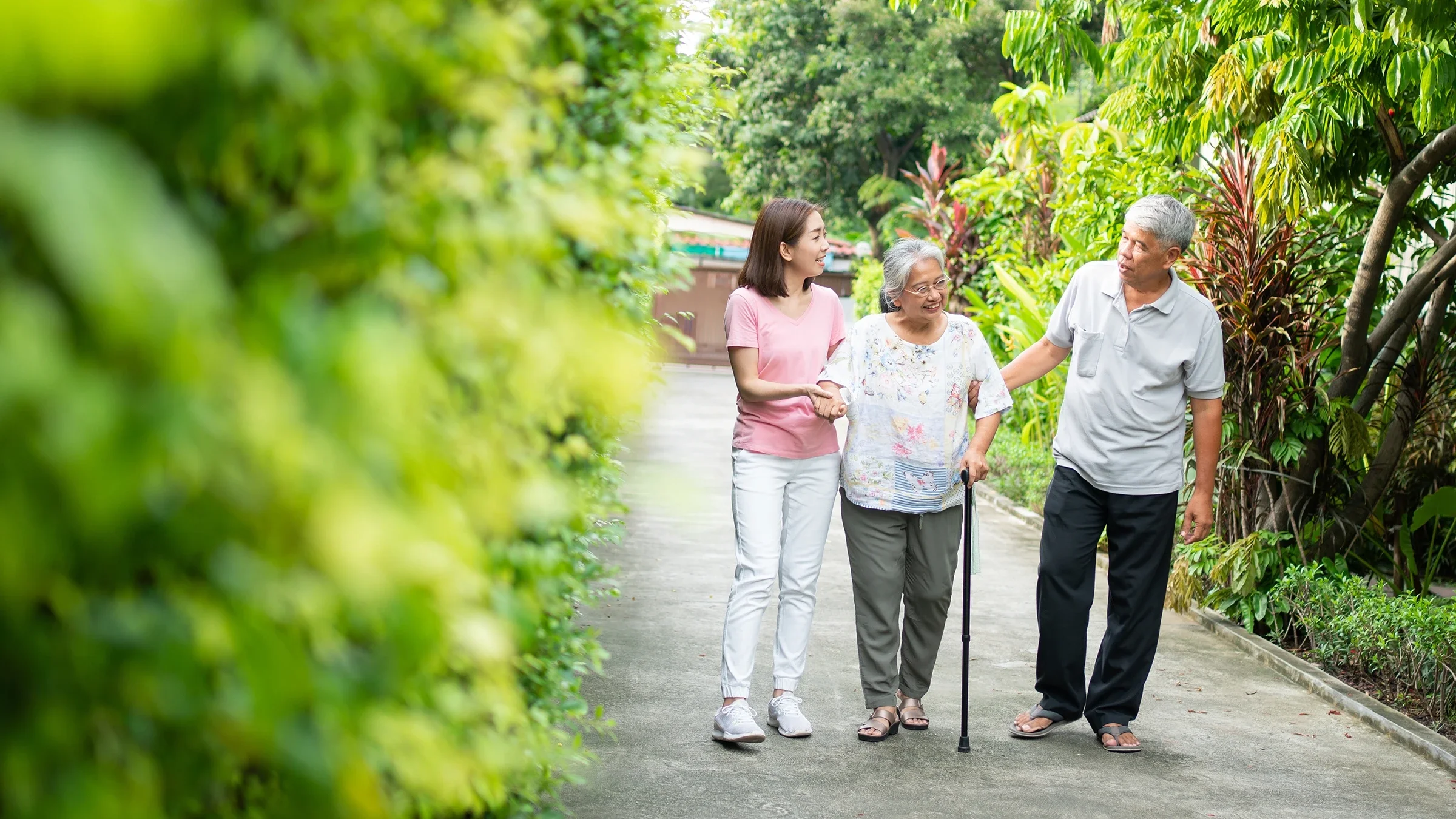 Adult daughter helping her mother on a family walk down a lush and green pathway. The mother is using a cane to walk and the father is on the other side of her.