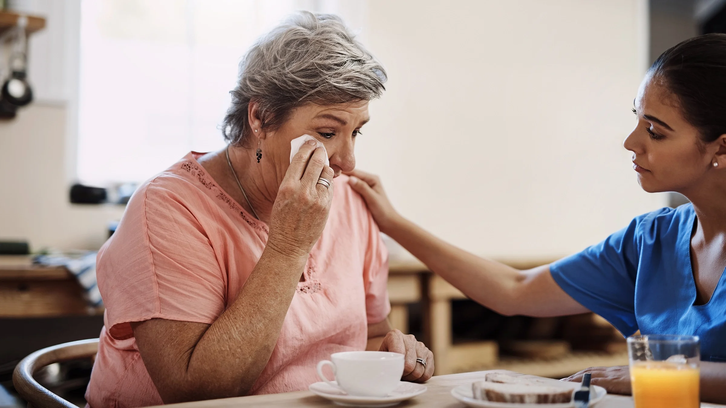 A female caregiver consoles a senior woman, who is crying.