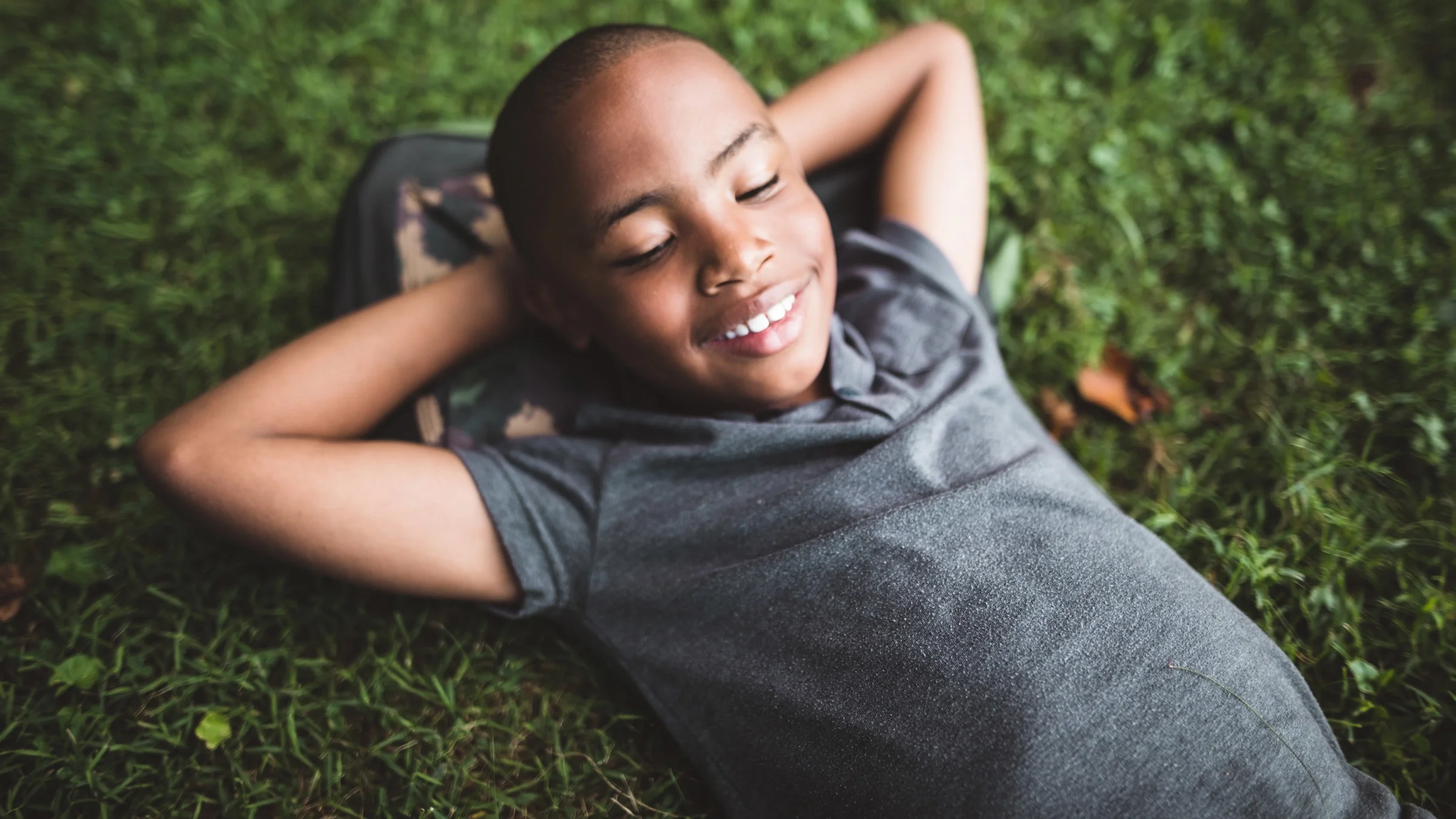 A young kid relaxing on the grass with his backpack as a pillow and hands behind his head.