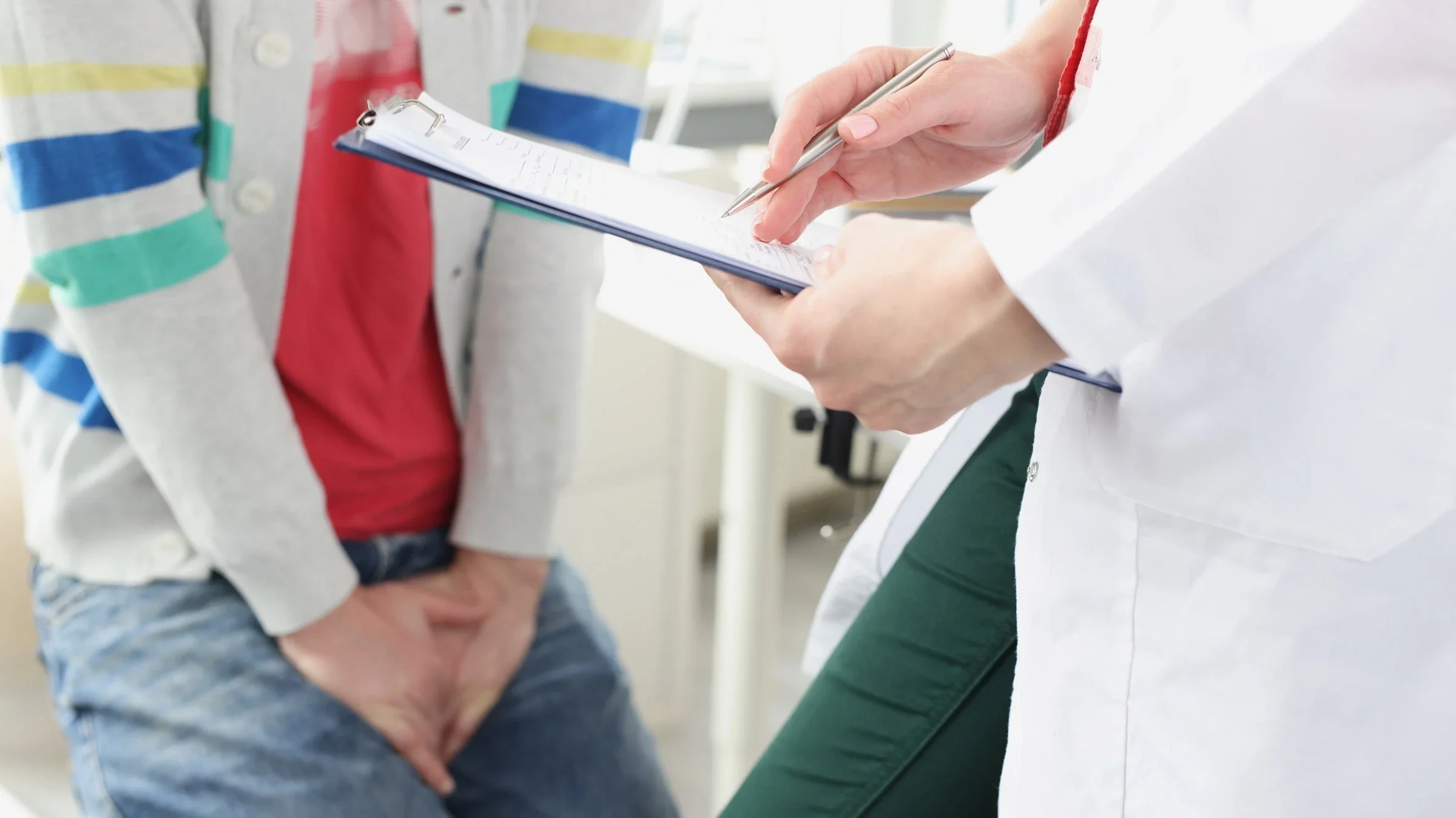 A cropped shot of a patient and a doctor in a urology appointment. The patient is holding their bladder and the patient is writing on a clipboard.