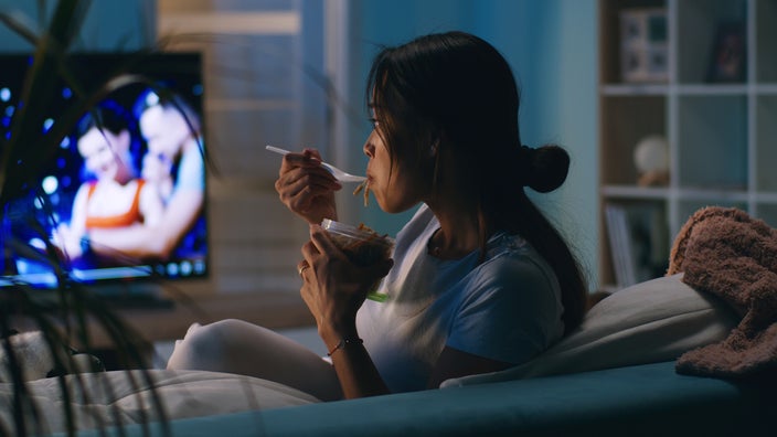 A woman watches TV while sitting on the couch and having dinner.
