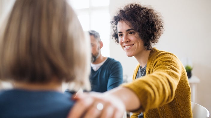 Support group. A young woman is putting her hand on a friend's shoulder and smiling softly at them.