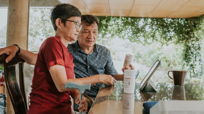 A elderly couple, male and female, looking at their tablet devise set on the dining table. They have different pill bottles in their hands.