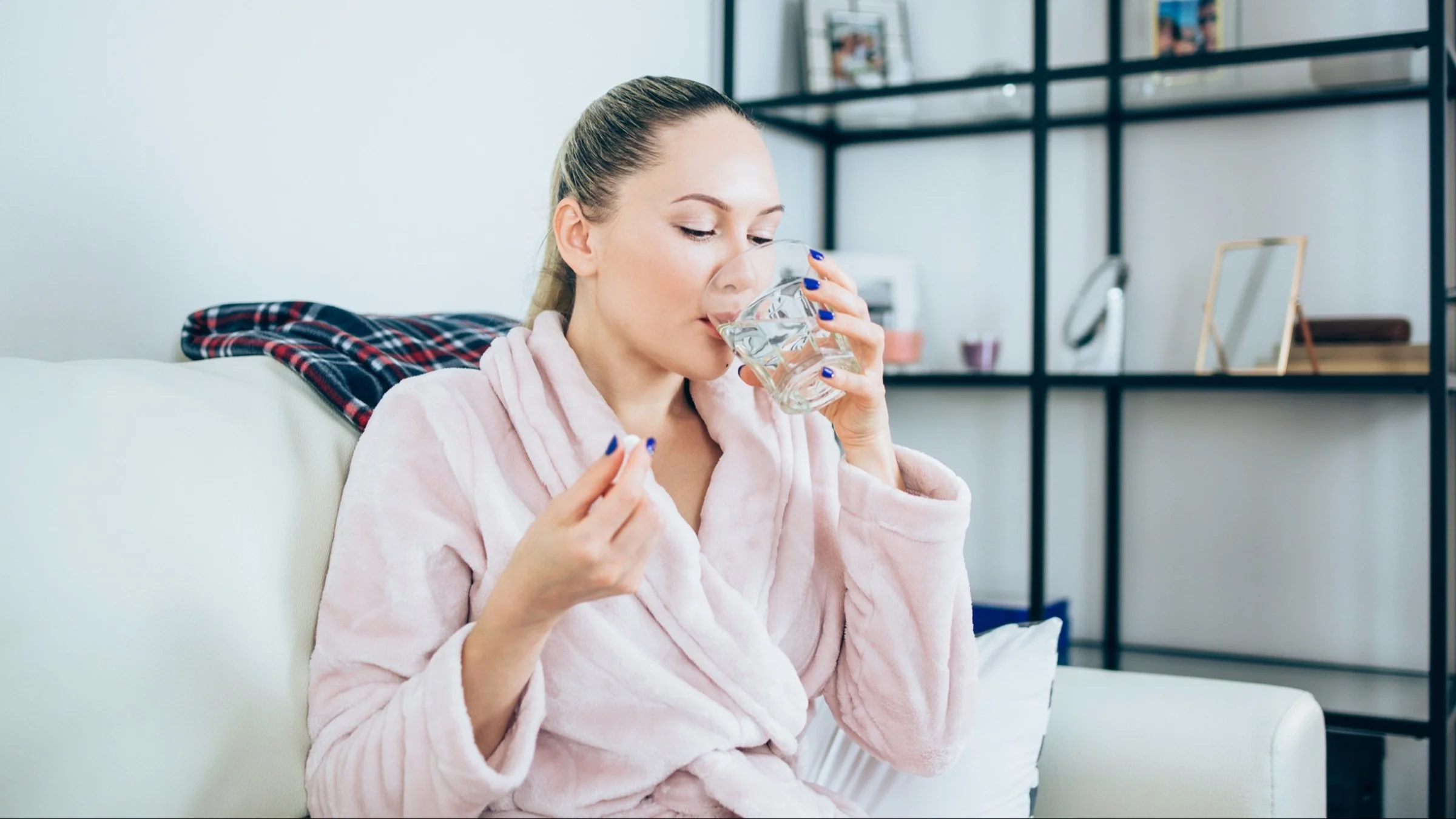Woman in pink robe sitting on the couch taking her antibiotics with a glass of water.