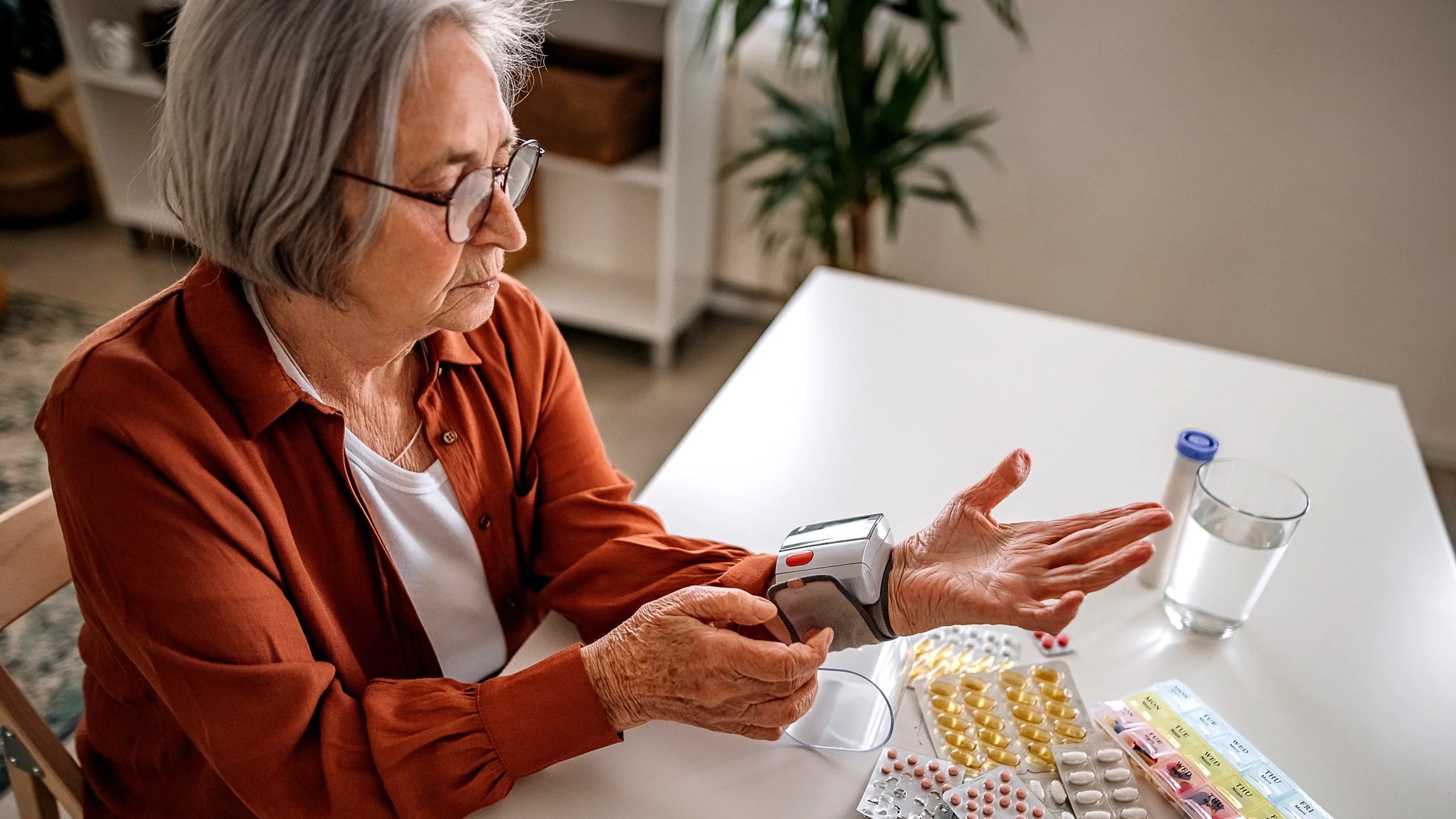 Senior woman taking her vitals with a blood pressure cuff at her kitchen table with pills surrounding her.