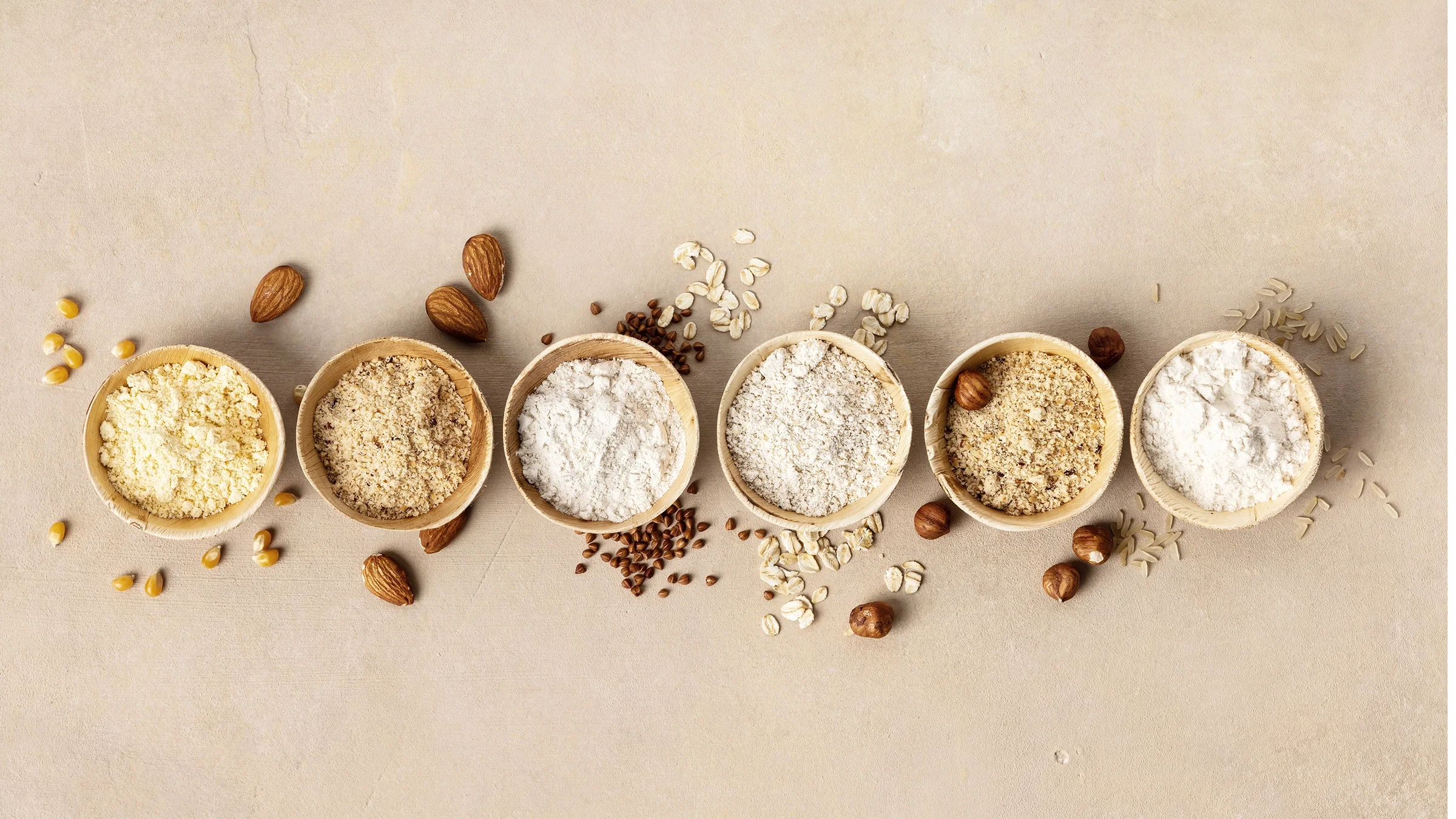 There are six bowls of various kinds of gluten-free flour with some sprinkled out on a tan background.