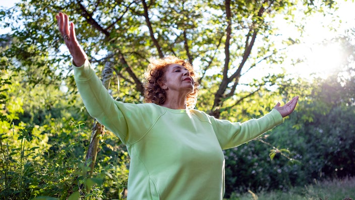 A woman stands outside in the sunshine. Her arms are extended out and her eyes are closed.