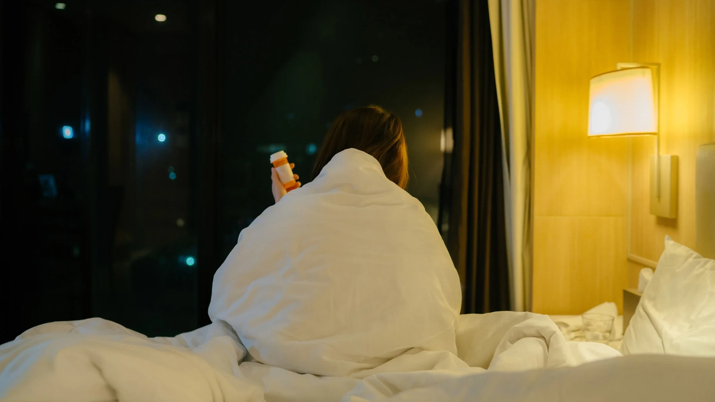 A person holding a pill bottle and shivering under their covers at night. 