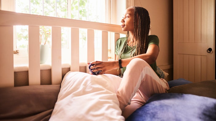 Young woman with braids looking out her window while drinking a cup of tea. She is wearing a green t-shirt and sweatpants.