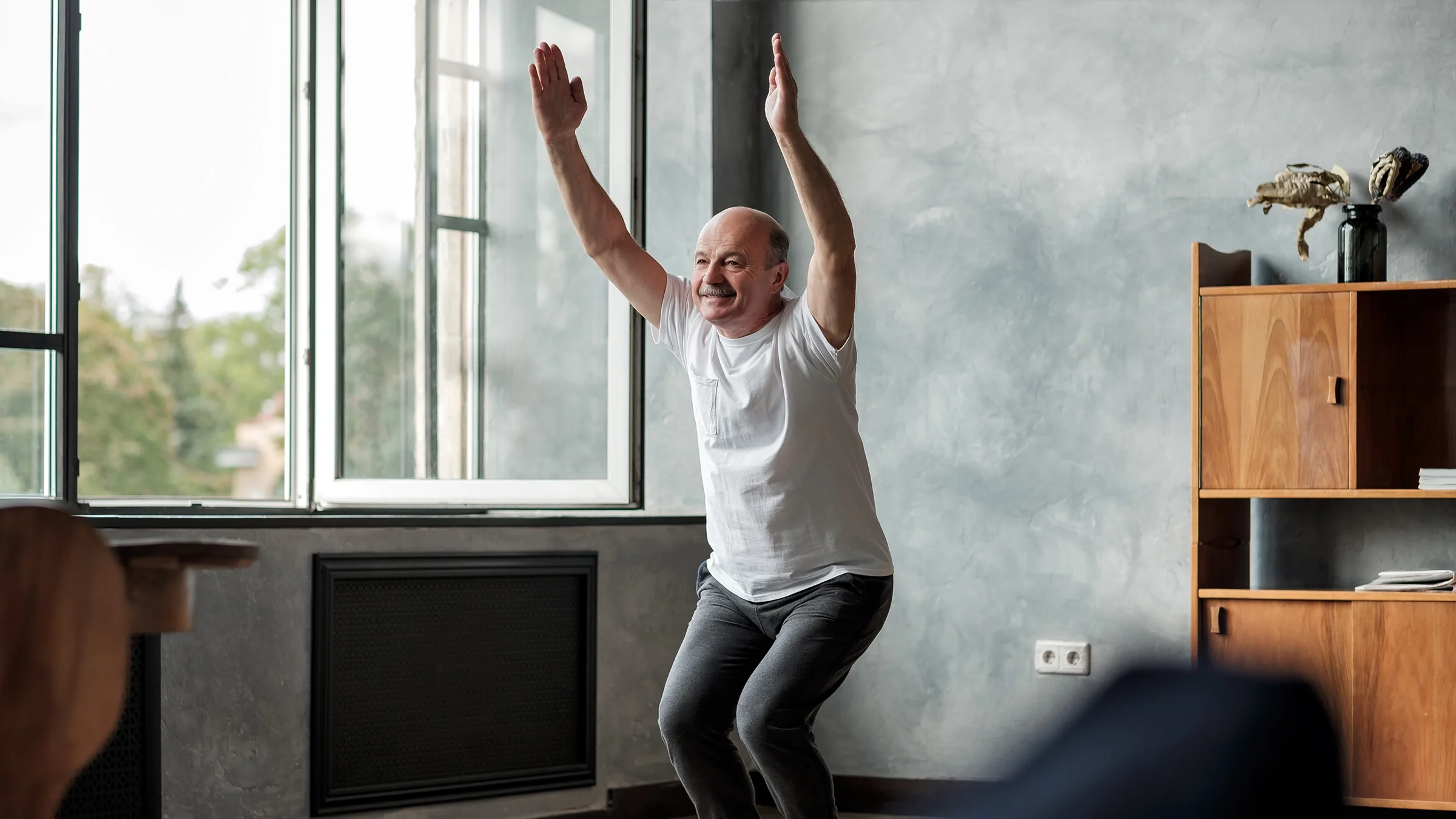 An older man is doing a yoga chair pose.