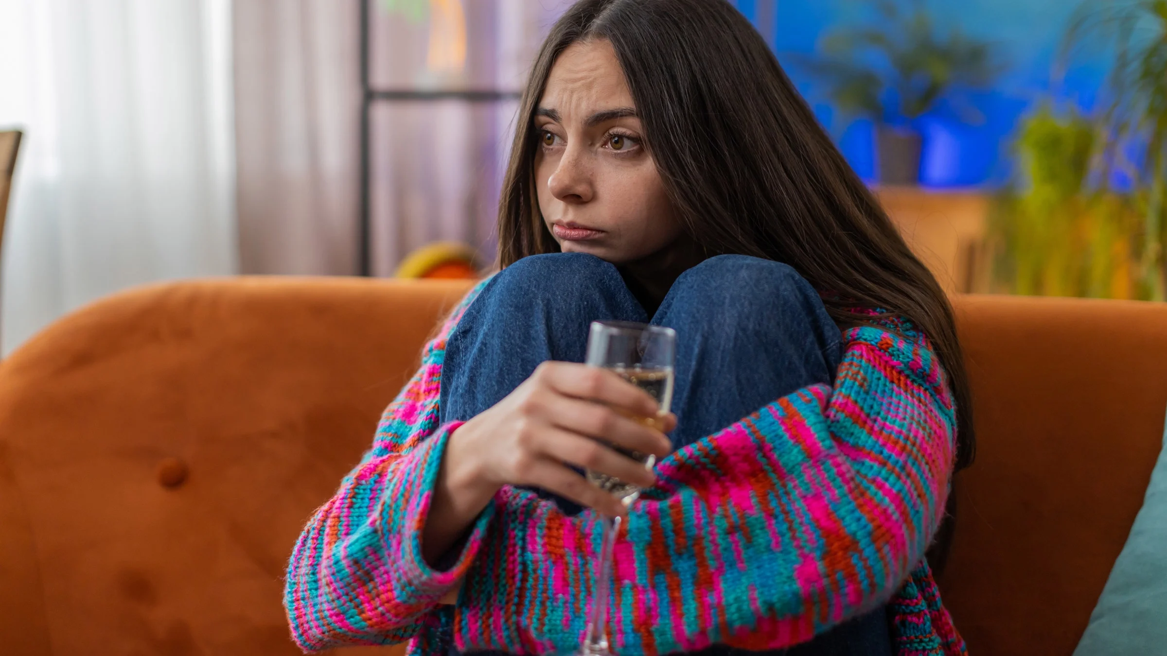 A person sitting on a couch holding a glass of alcohol with a nervous expression.