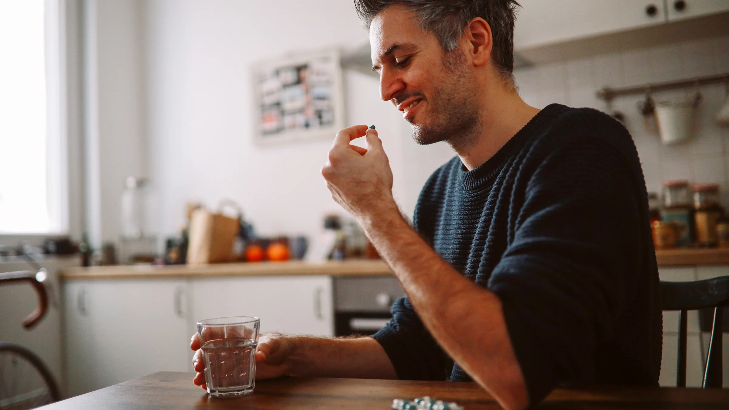 A man sitting at a kitchen table takes a pill.
